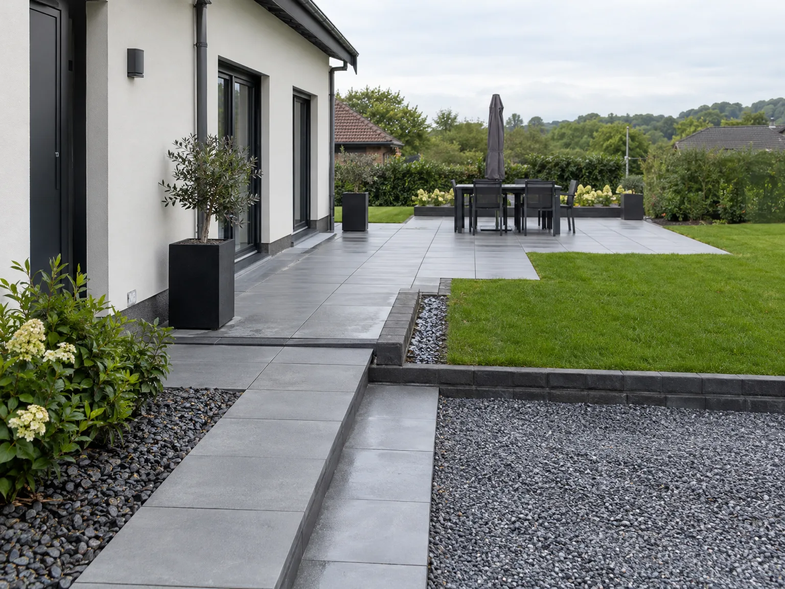 Terrasse moderne dallée grise avec salon de jardin noir, fleurs et gazon verdoyant